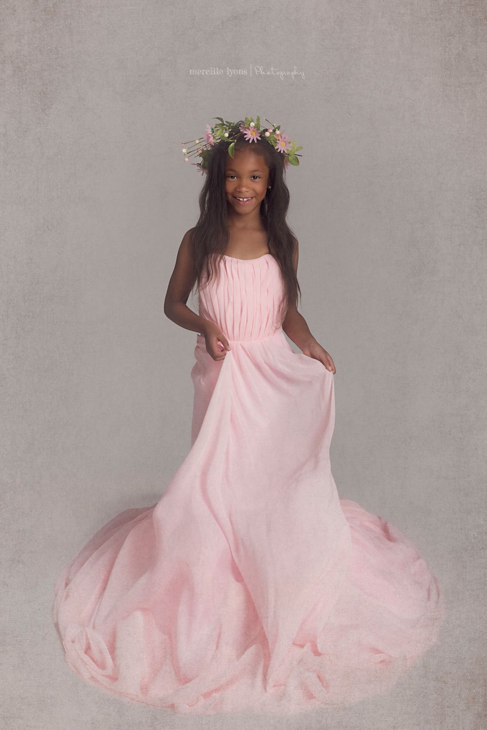 Young dancer wearing pink flowing gown and floral crown poses elegantly.