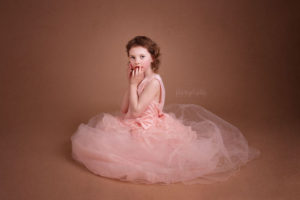 Young dancer in pink tulle dress poses gracefully against brown backdrop.