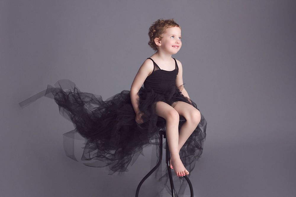 Young performer in black tulle dress sits elegantly on stool against grey backdrop.