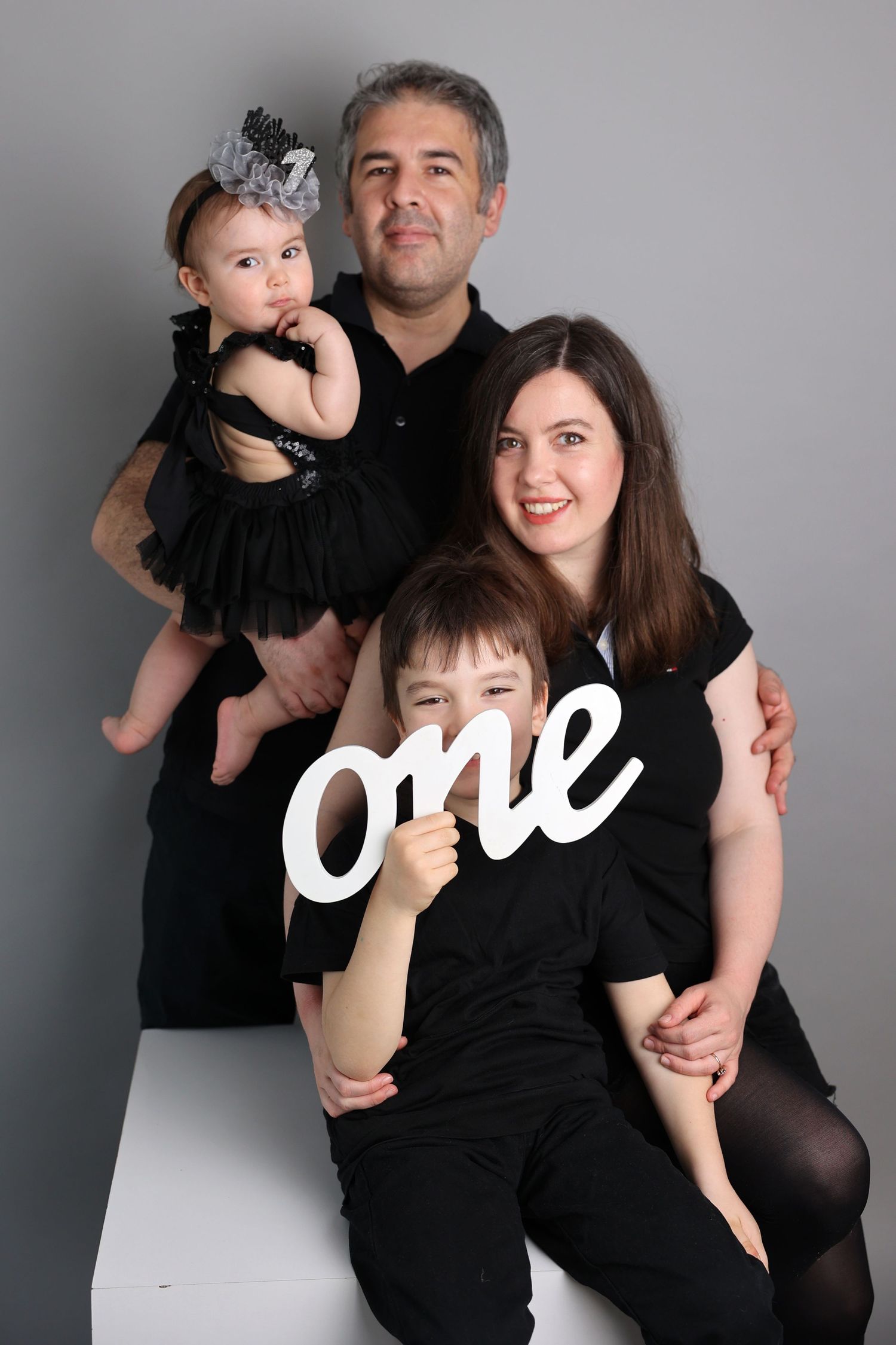 Family portrait featuring a white 'one' prop with parents and children in black attire against gray background.