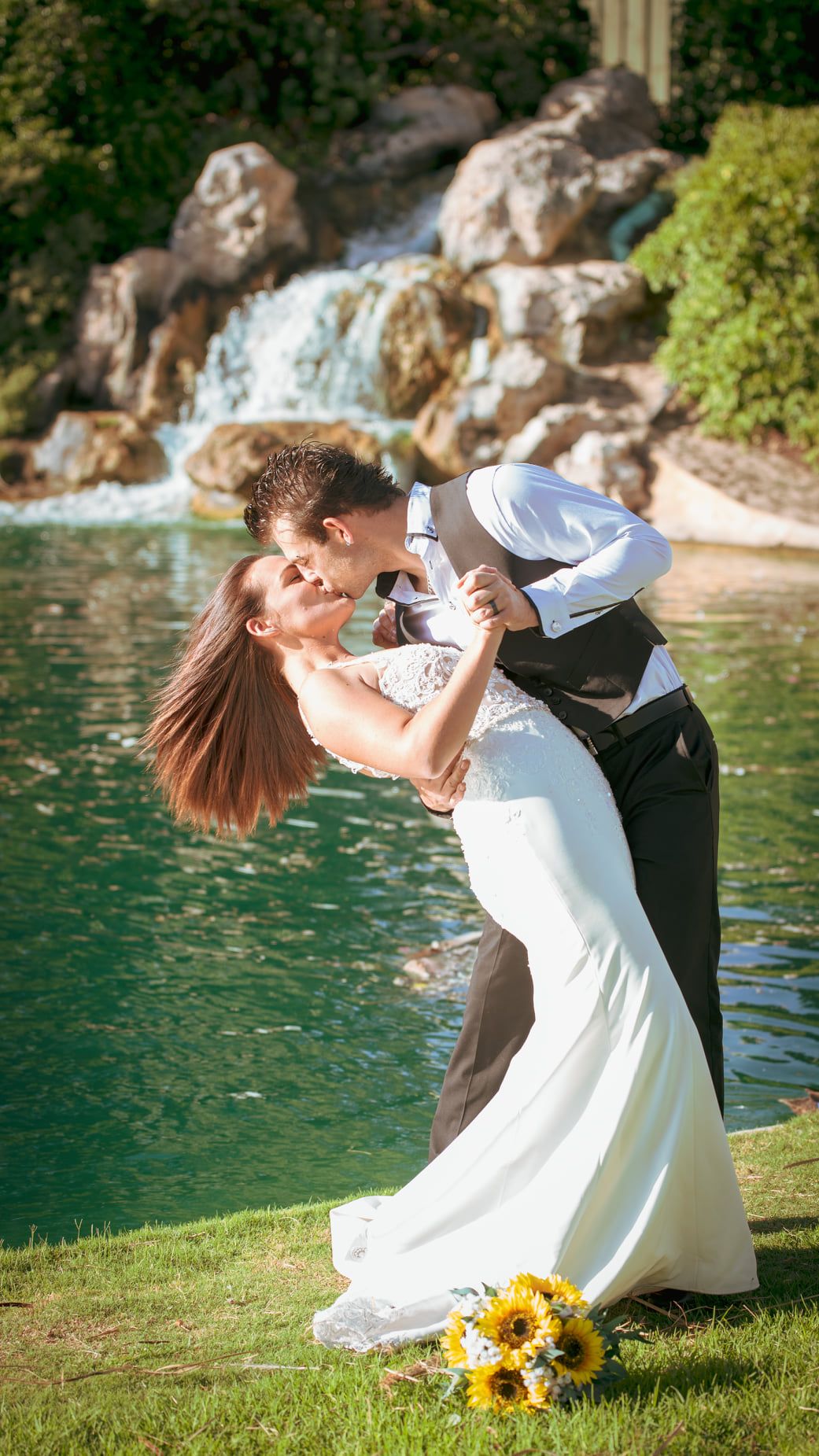 A romantic wedding photo showing a couple dancing by a waterfall with sunflowers on the ground.