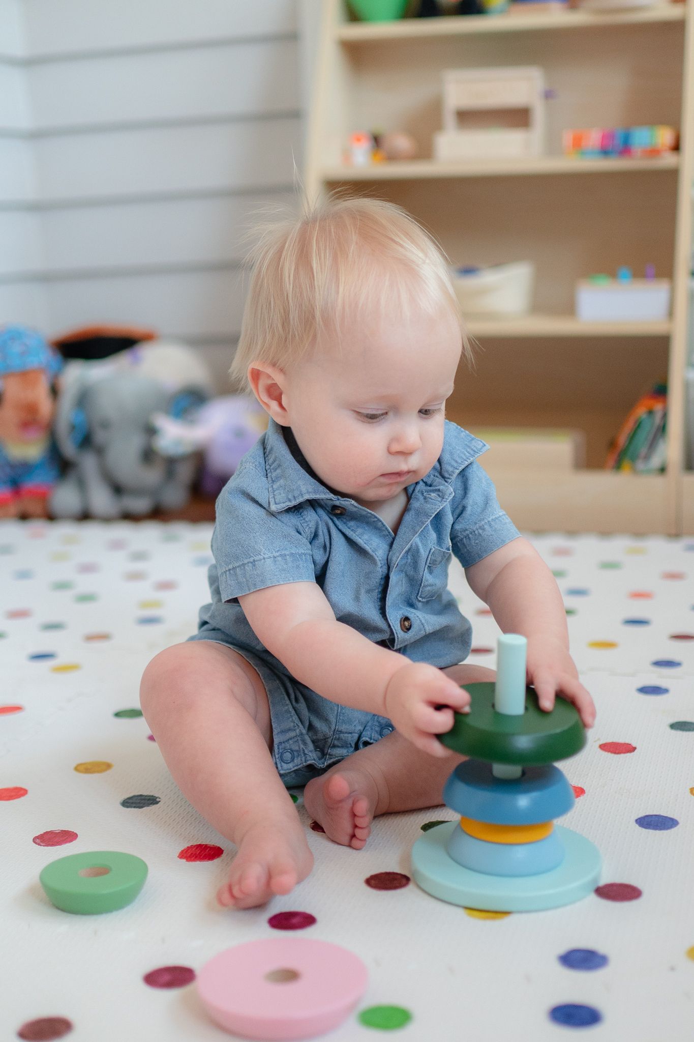 A toddler in blue plays with a colorful stacking toy on a polka dot mat.