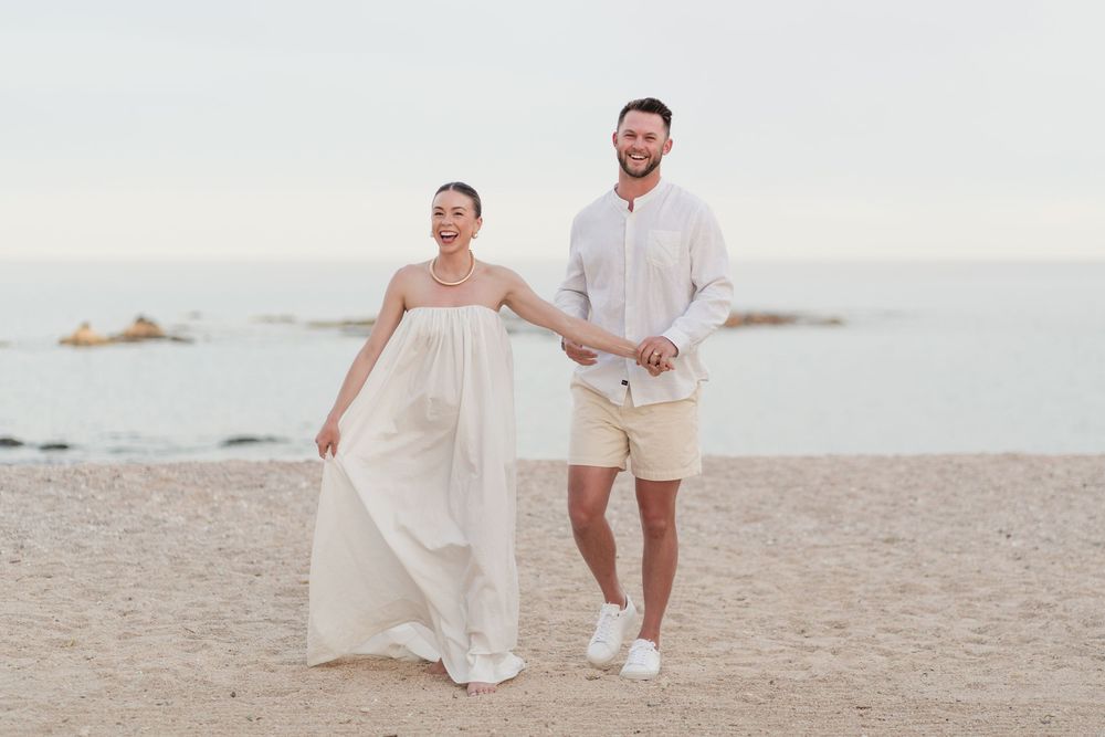 Happy couple in light-colored attire walks playfully along a scenic beach during sunset.