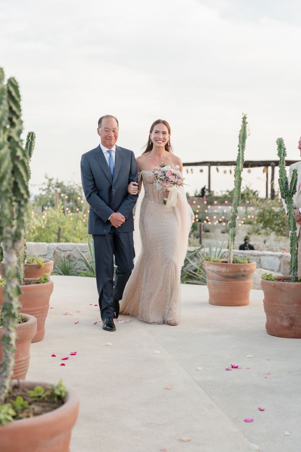 An outdoor wedding scene with couple walking down a paved aisle lined with potted desert plants and rose petals.