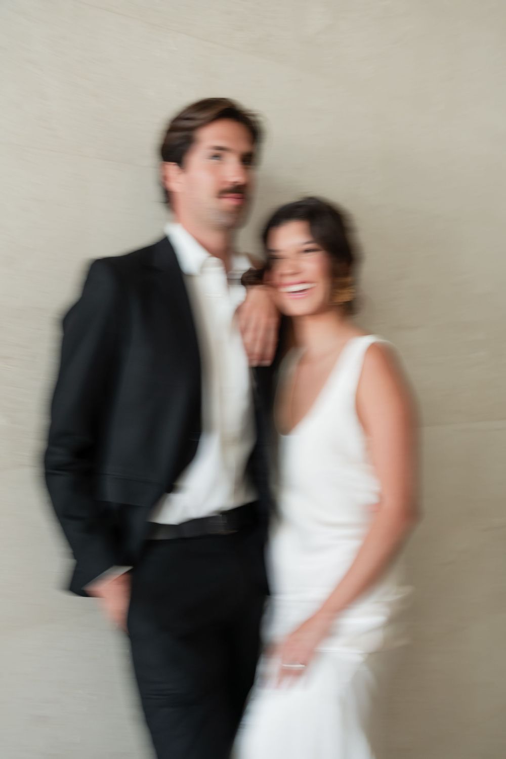 Motion-blurred wedding portrait of a couple in elegant black suit and white dress smiling against a light wall.