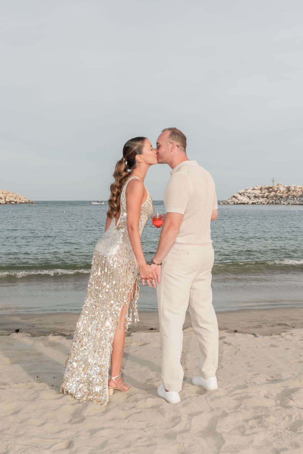 A romantic beach wedding couple sharing a kiss at sunset in elegant cream and sparkly attire.