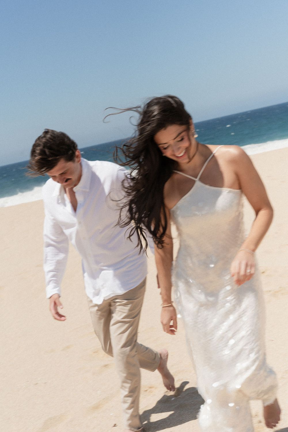Happy couple in white attire running barefoot on a sunny beach with ocean waves in the background.
