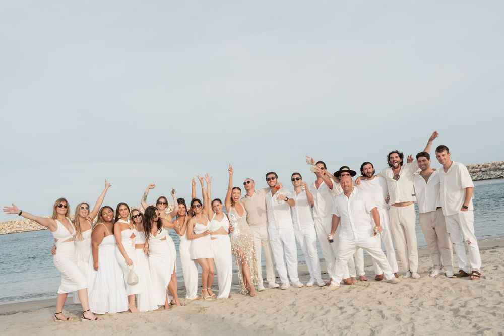 A large wedding party dressed in white poses for photos along a scenic beach at sunset.
