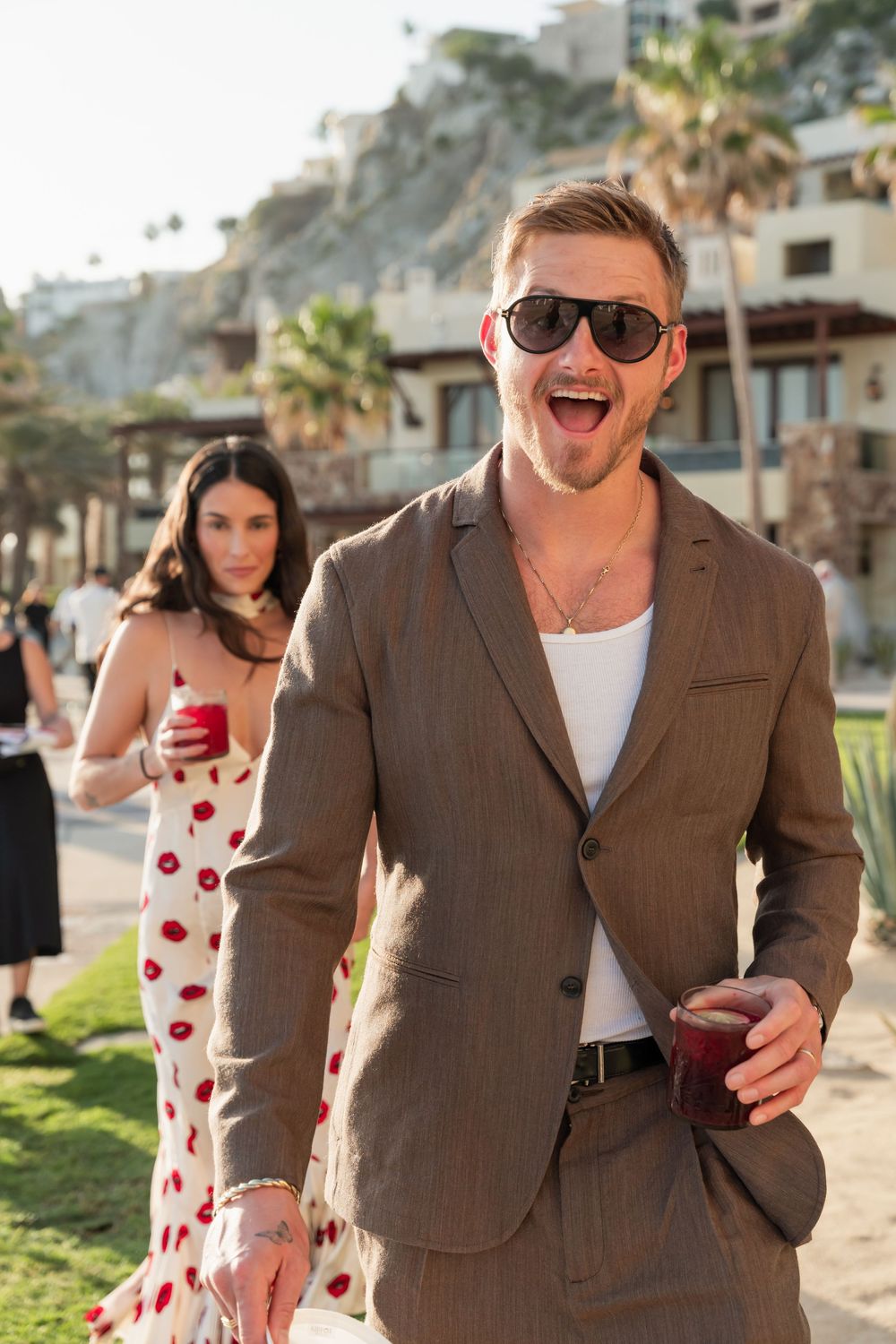 Person in brown blazer and sunglasses laughing while someone in polka dot dress walks behind at a beachside venue.