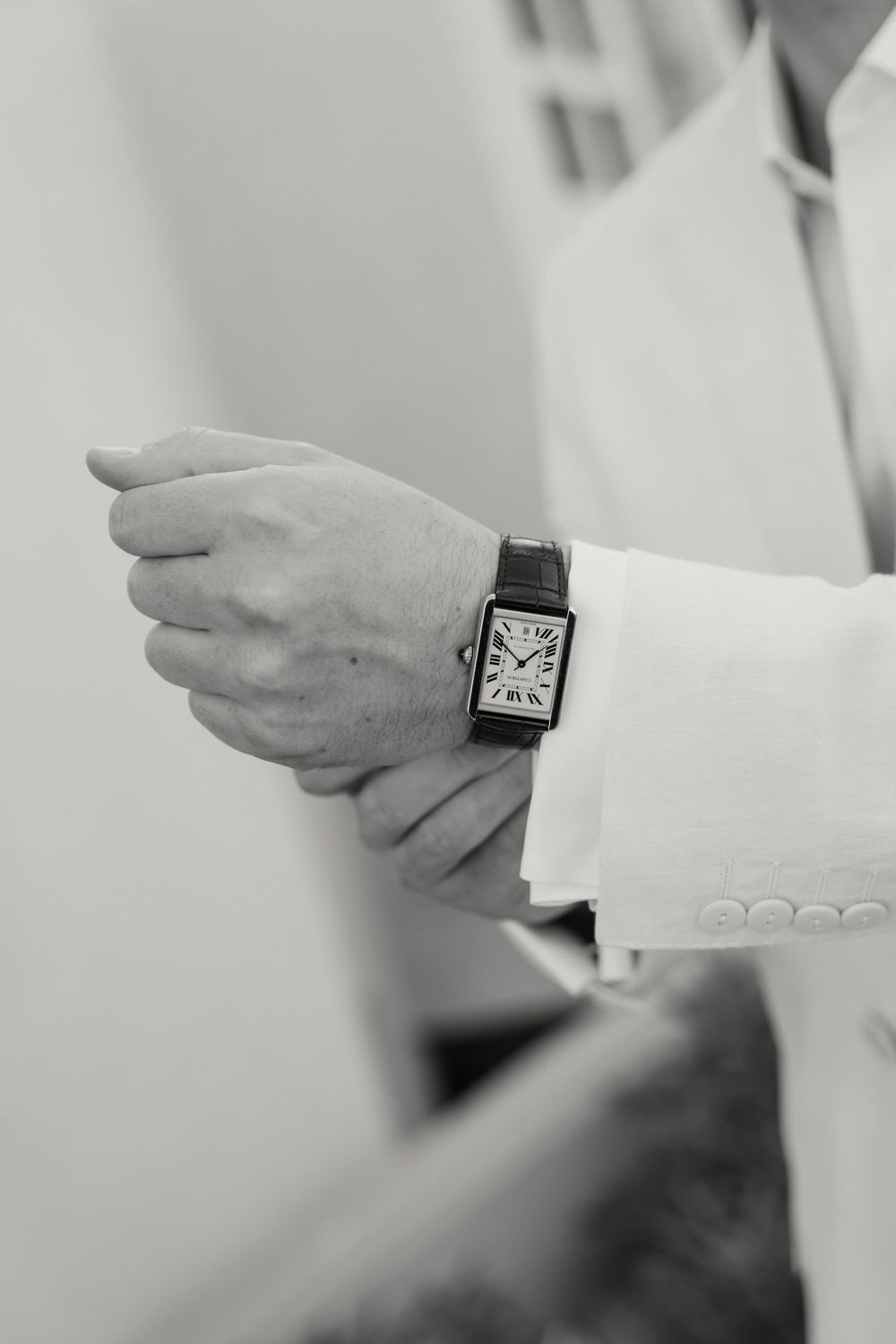 Black and white detail shot shows an elegant square wristwatch worn with a light colored suit sleeve.