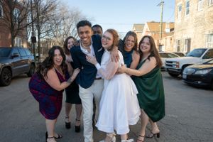 A group hug photo of a newlywed couple and their wedding party in the streets under a blue sky.