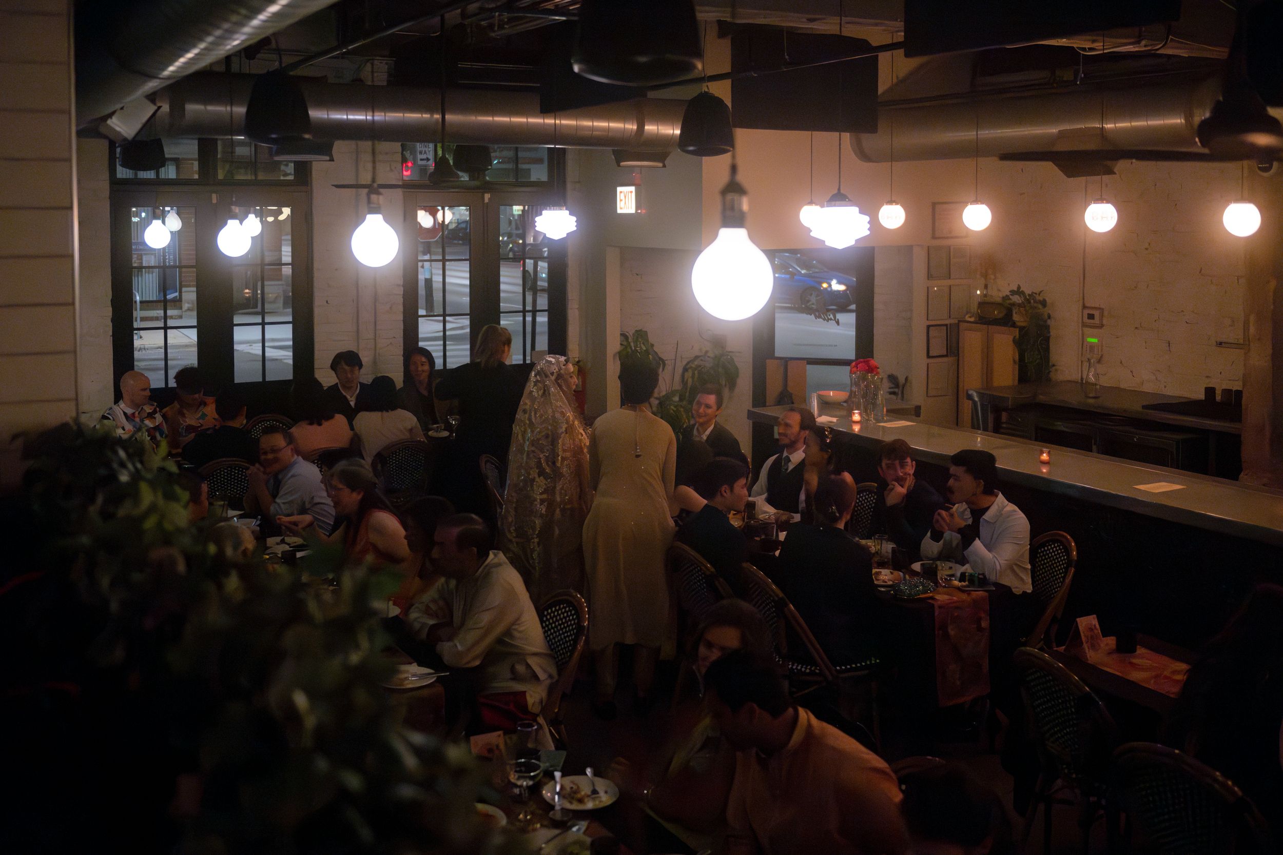 A queer, newlywed couple greets their guests at dinner, in between two long tables.