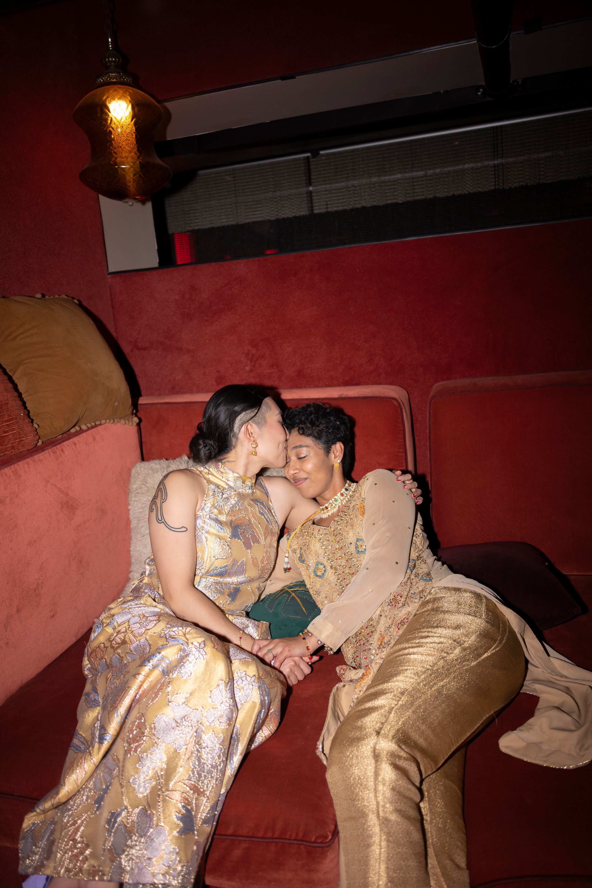 A queer, interracial couple reclines on velvety seating during their reception, and one kisses the other on the temple.