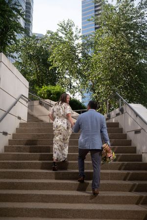 A groom follows his bride up outdoor stairs. He holds her bouquet in one hand and her hand in the other, she looks back at him as they climb