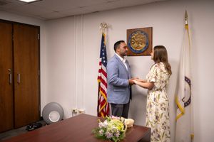 Bouquet on the table, a couple stands facing each other, hands held in the middle, flanked by the US and Illinois flags, at City Hall.