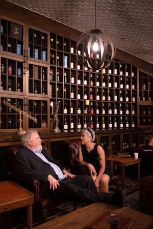 Two wedding guests sit and converse in a cellar lounge with bottles of wine on shelves lining the wall in the background.