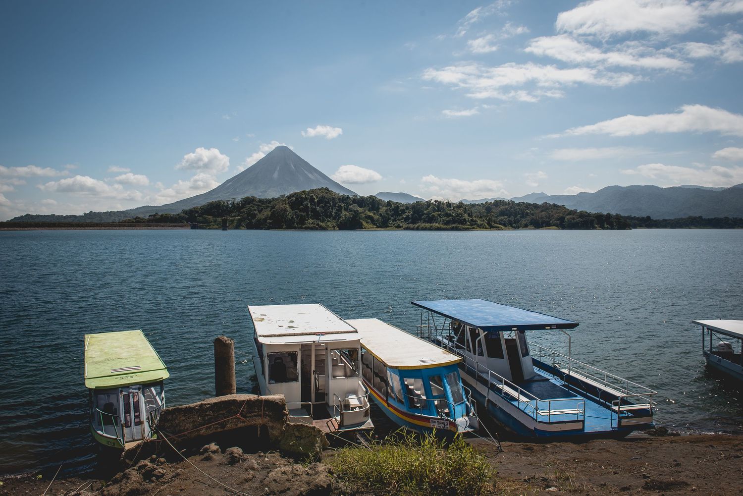 Boats docked on a lake with a majestic volcano mountain peak in the background under blue skies.