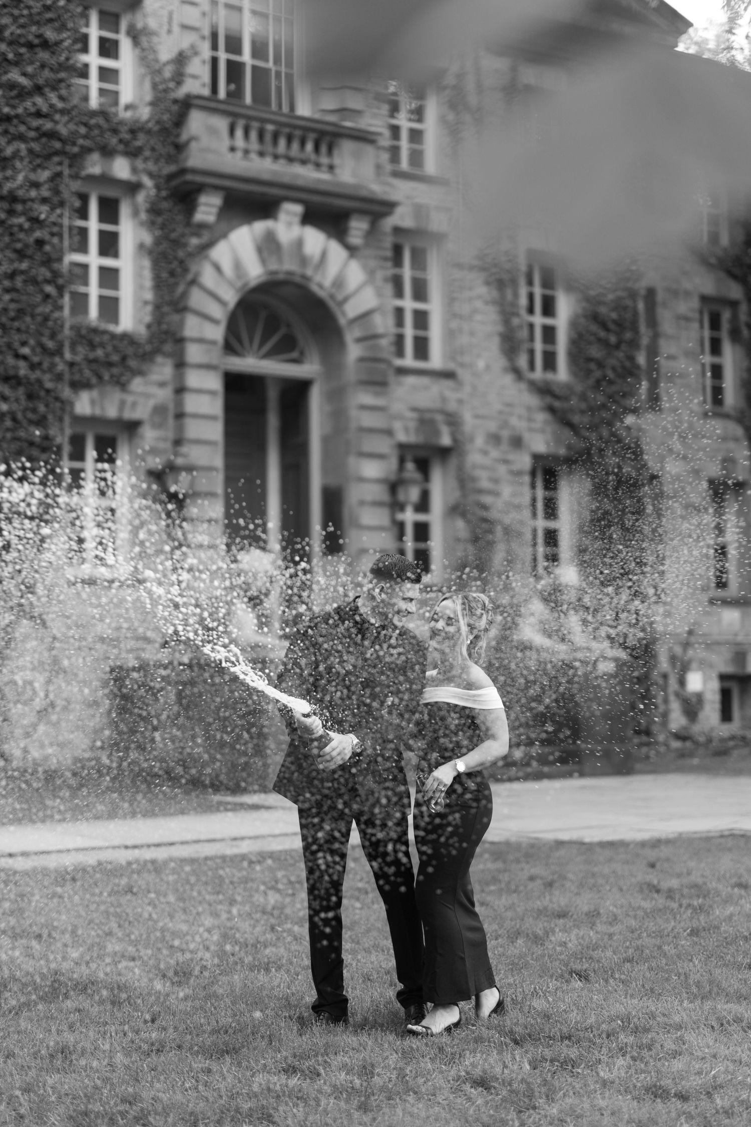 Black and white photo of a couple popping champagne with bubbles spraying before an ivy-covered mansion.