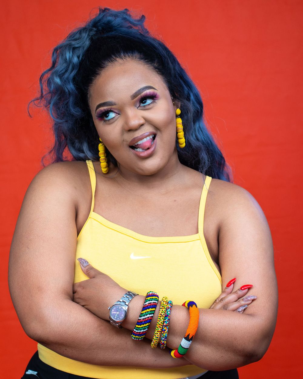 Playful portrait with blue hair wearing yellow top and colorful bracelets against red background.
