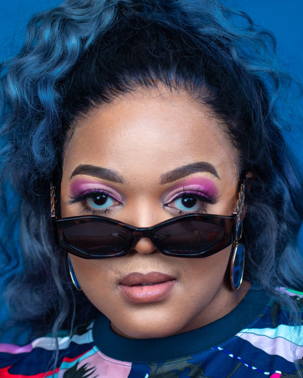 Fashion portrait with vibrant blue hair, pink eyeshadow and dark sunglasses against blue background.