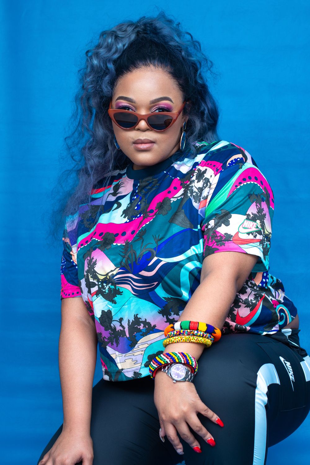 Fashion portrait with colorful patterned shirt, sunglasses, and accessories against blue background.