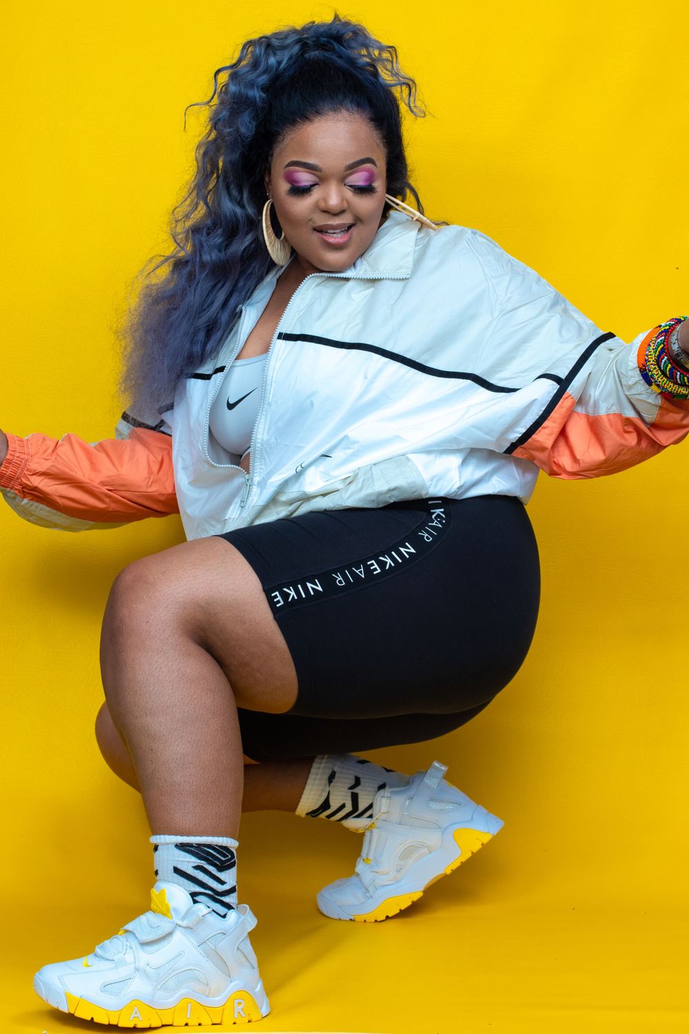 Athletic fashion pose in Nike windbreaker and shorts against bright yellow background.
