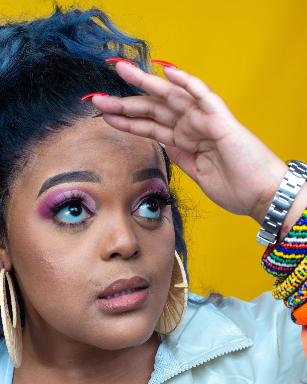 Portrait against yellow background featuring vibrant blue hair, colorful makeup and stacked bracelets.