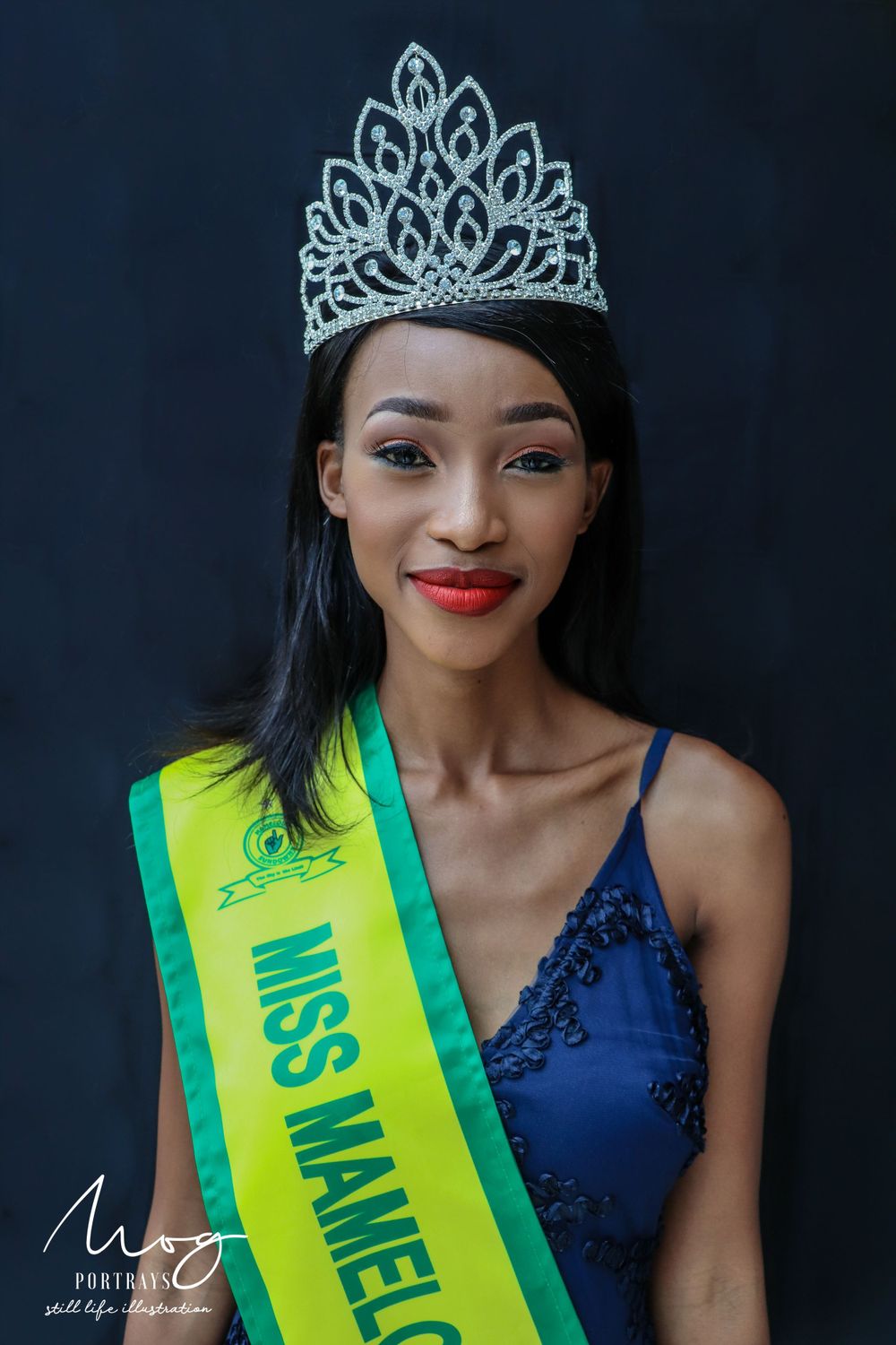 Beauty pageant contestant wearing a sparkling crown and yellow-green sash against dark background.