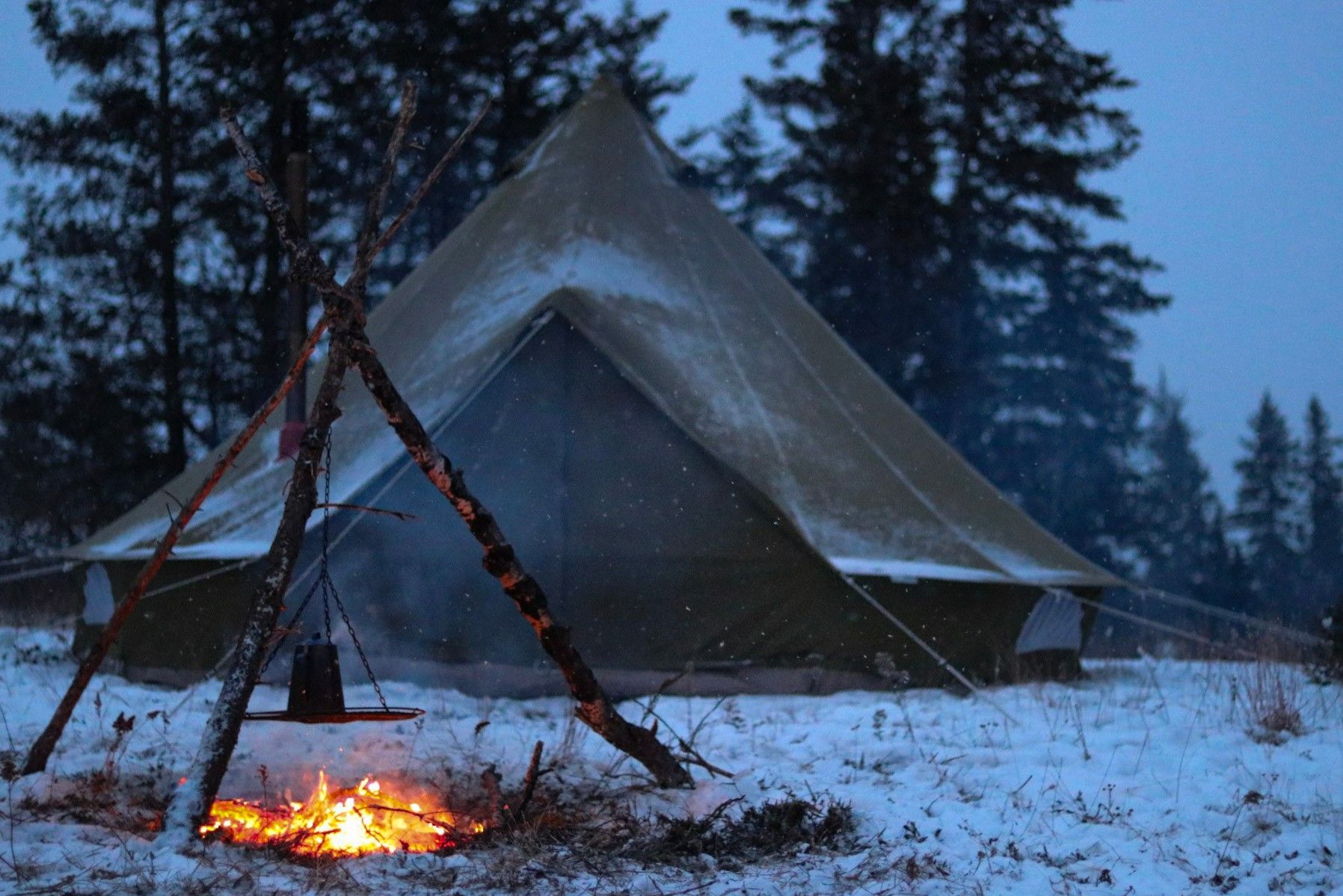 Snow-covered bell tent with campfire glowing at dusk in winter forest.