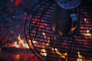 Close-up of food cooking on round grill grate over glowing campfire embers.