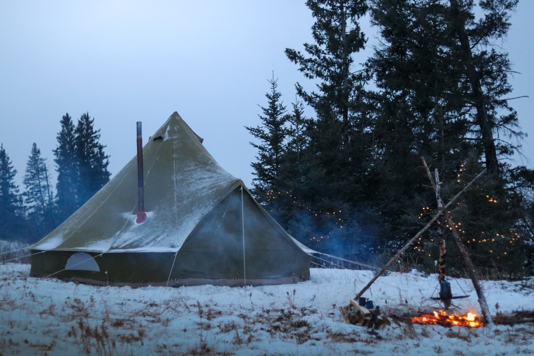Canvas tent sits in snowy forest at dusk with campfire and string lights creating cozy winter ambiance.