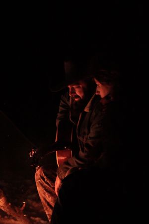 Two figures sit in the darkness illuminated by a warm orange glow from a campfire at night.