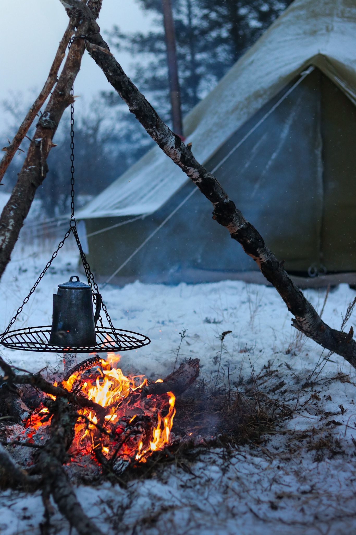 A metal kettle hangs over a glowing campfire beside a snow-covered tent in winter.