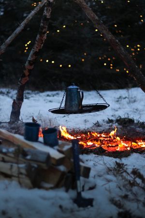 Coffee pot suspended over campfire with string lights twinkling in snowy winter background.