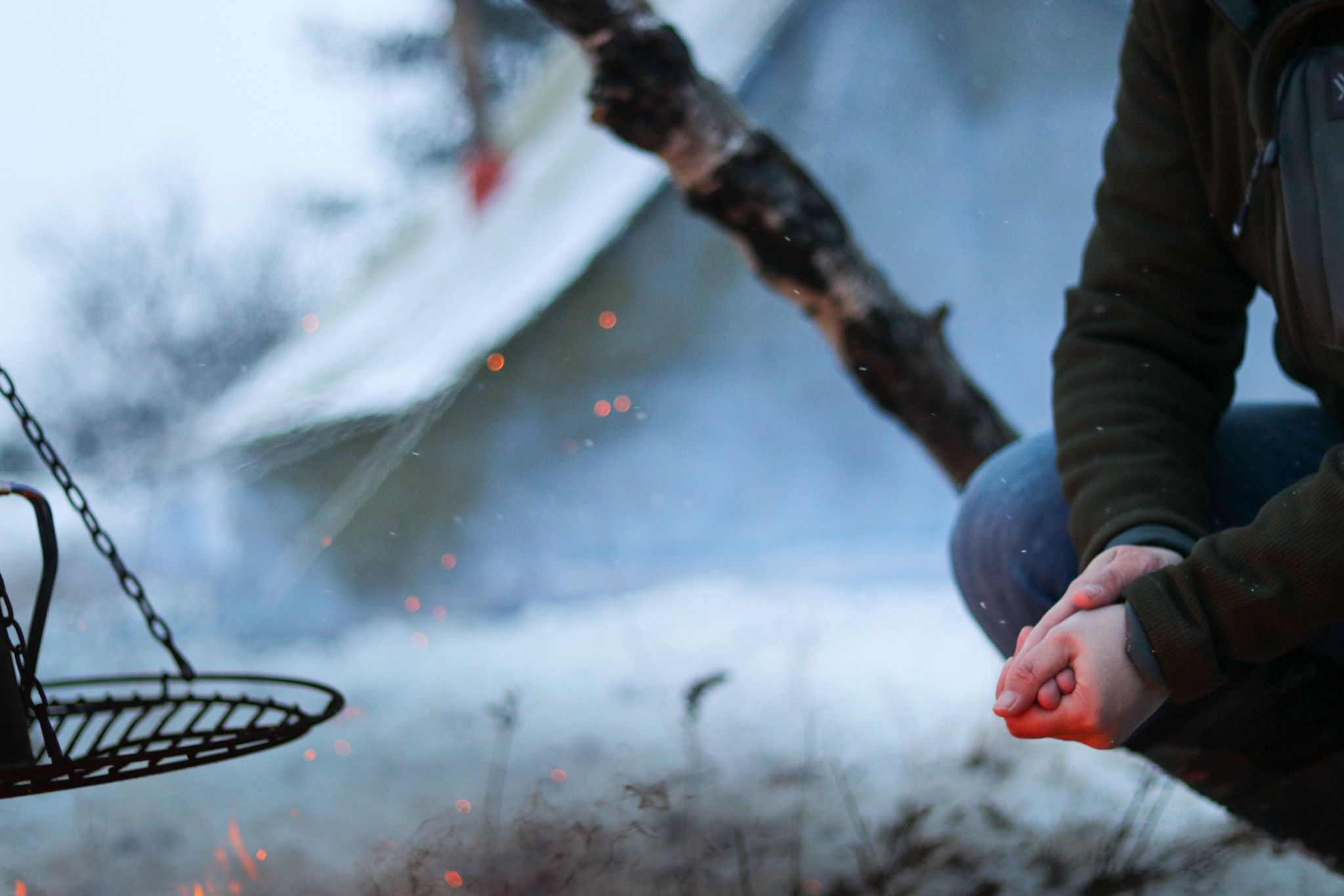 Hands warming by a winter campfire with suspended cooking grate in the foreground.