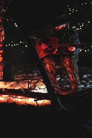 Someone playing acoustic guitar by a campfire at night with string lights in the background.