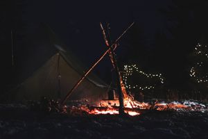 A glowing campfire illuminates a tent in the snow with twinkling lights in the distance.
