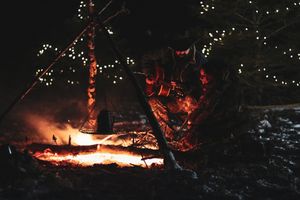 People gather around a bright campfire at night with string lights decorating the trees.