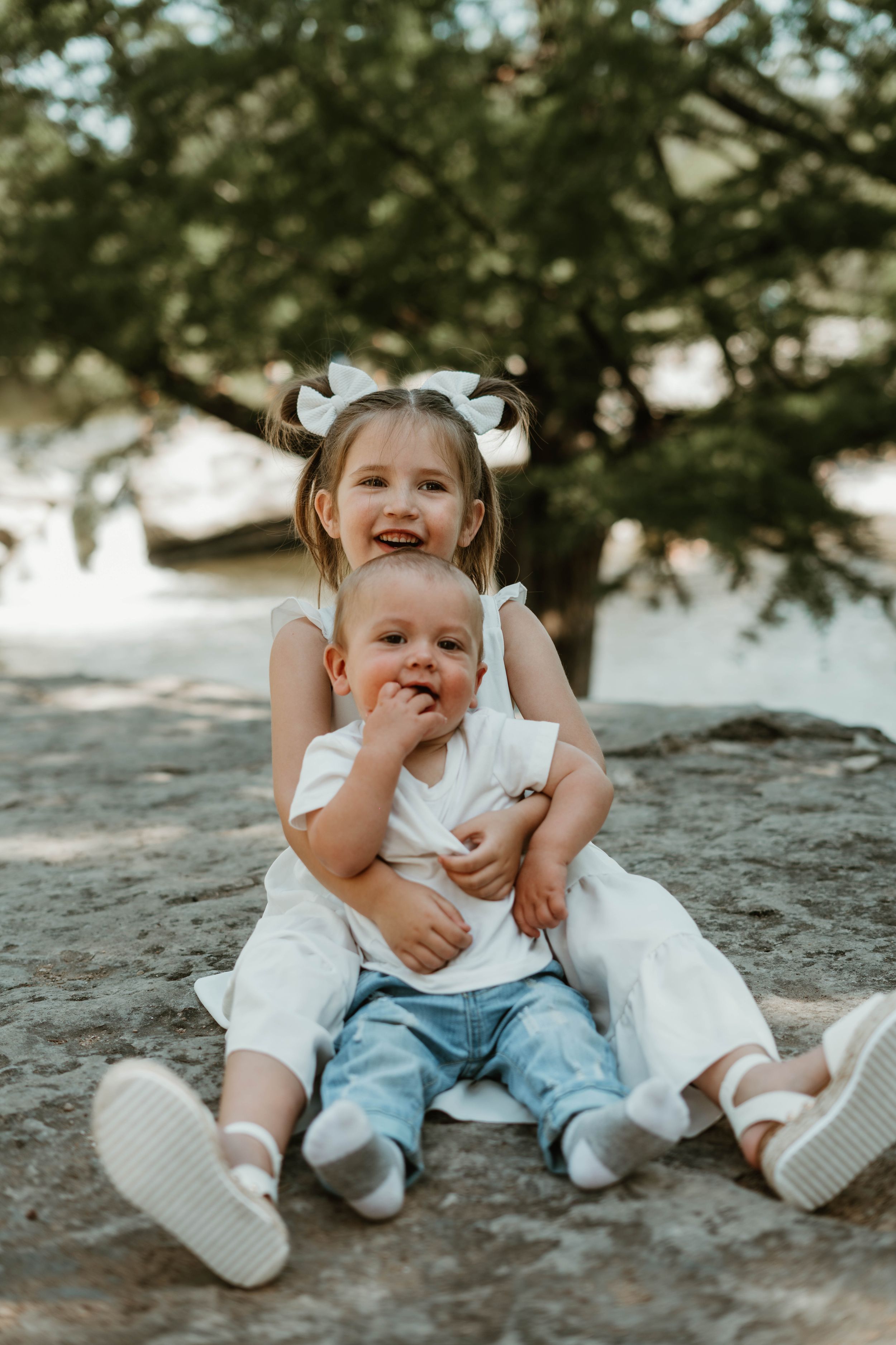 Two children in white outfits share a playful moment while sitting outdoors.