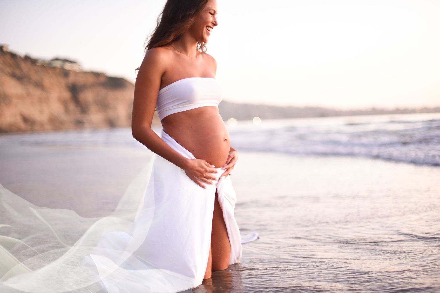 Maternity photos of woman in white dress on beach at sunset highlighting her pregnant belly.