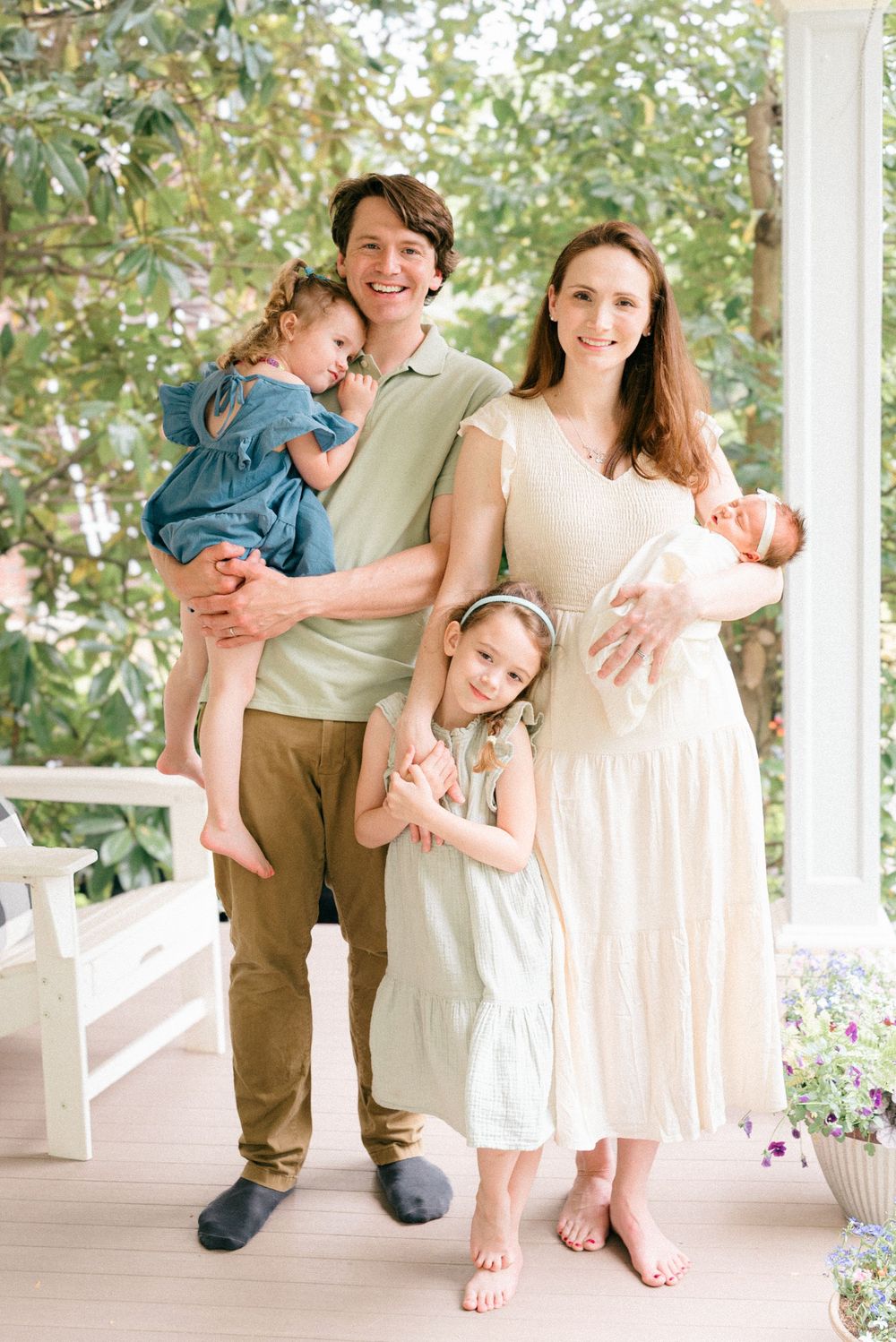 Intimate in-home motherhood photo of mother holding newborn and husband and other children pose in soft natural light on a porch.