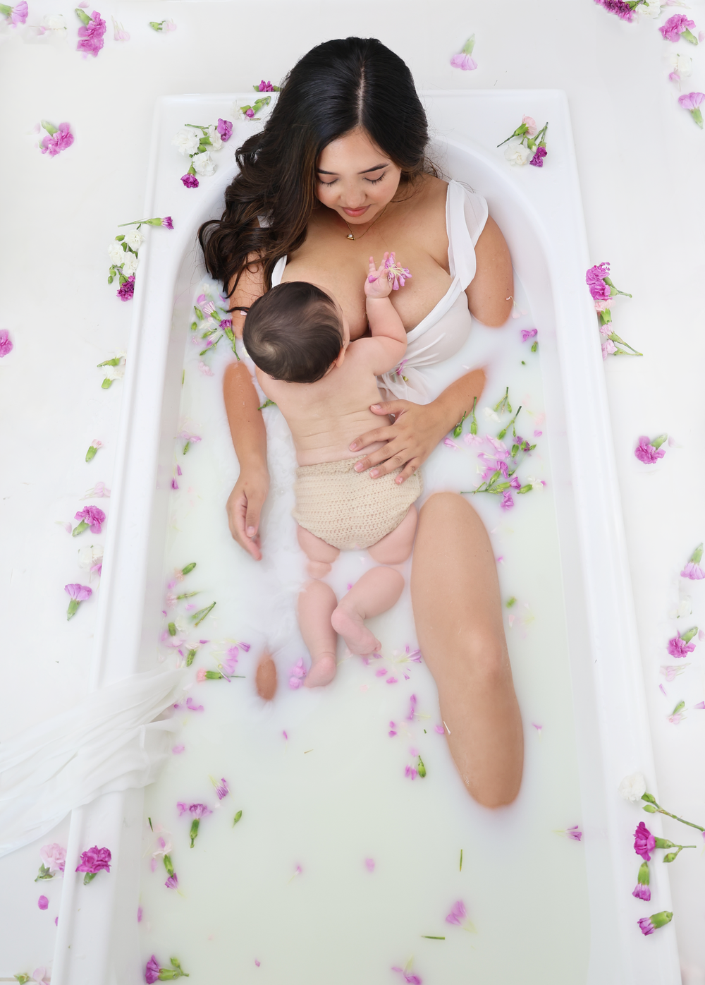 A milk bath maternity photoshoot with scattered pink flower petals and greenery on white background.
