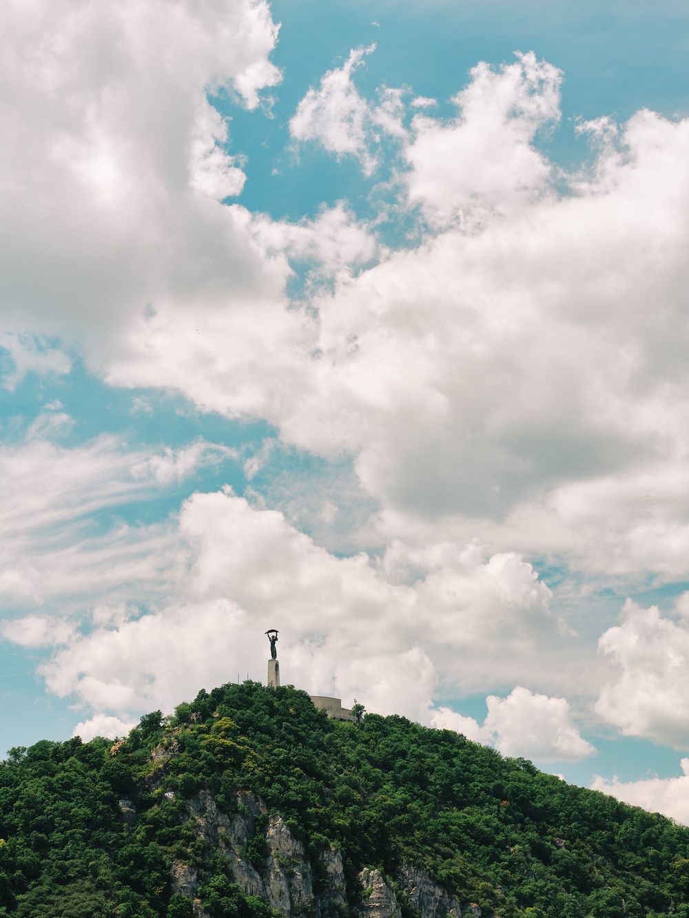 A tall monument stands atop a green forested hill against a dramatic cloudy blue sky.