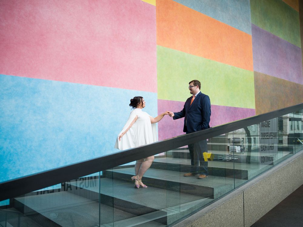 Two people in formal attire stand on stairs against a colorful geometric wall mural in an urban setting.