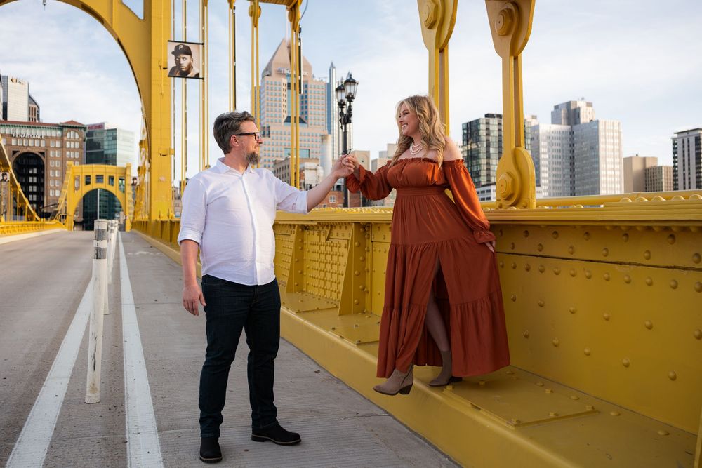 Two people share a candid moment while standing on Pittsburgh's iconic yellow Roberto Clemente Bridge with the city skyline behind.