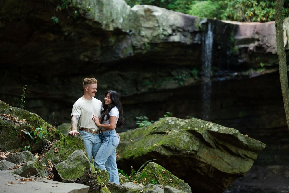 A romantic couple poses for photos across mossy cliffs and waterfall backdrops in a scenic outdoor engagement session.