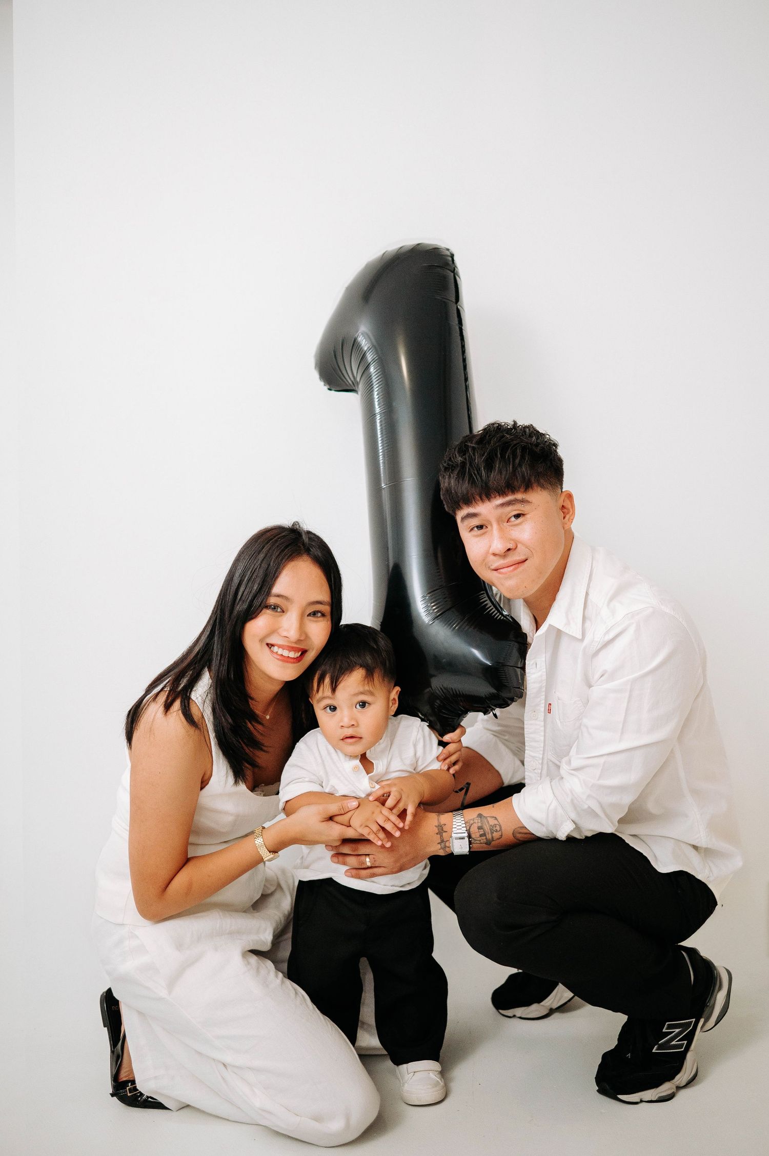 Family poses together in a minimalist white studio setting with a large black balloon number one decoration.