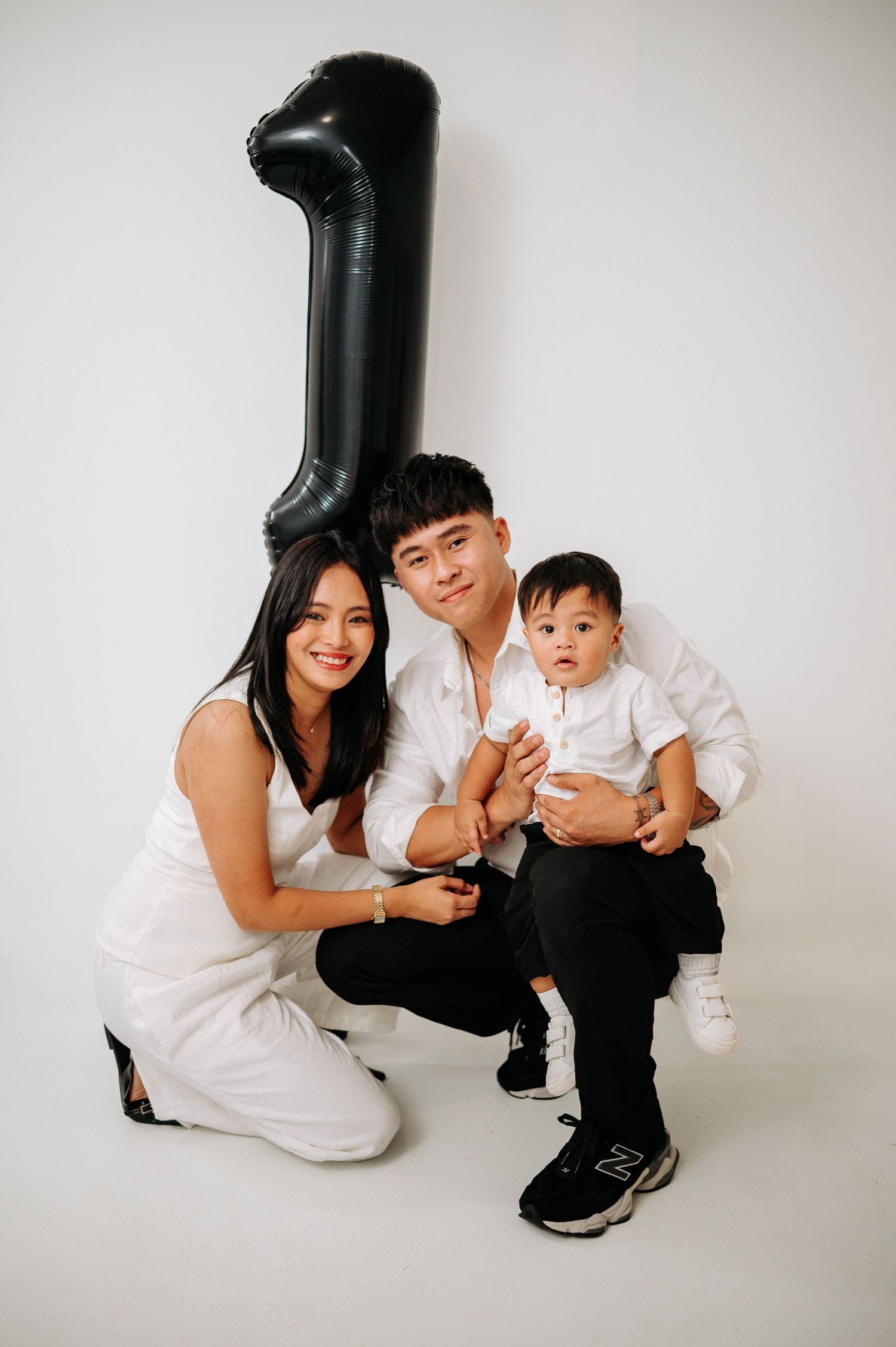 Family poses together with large black number one balloon against white backdrop during first birthday photoshoot.