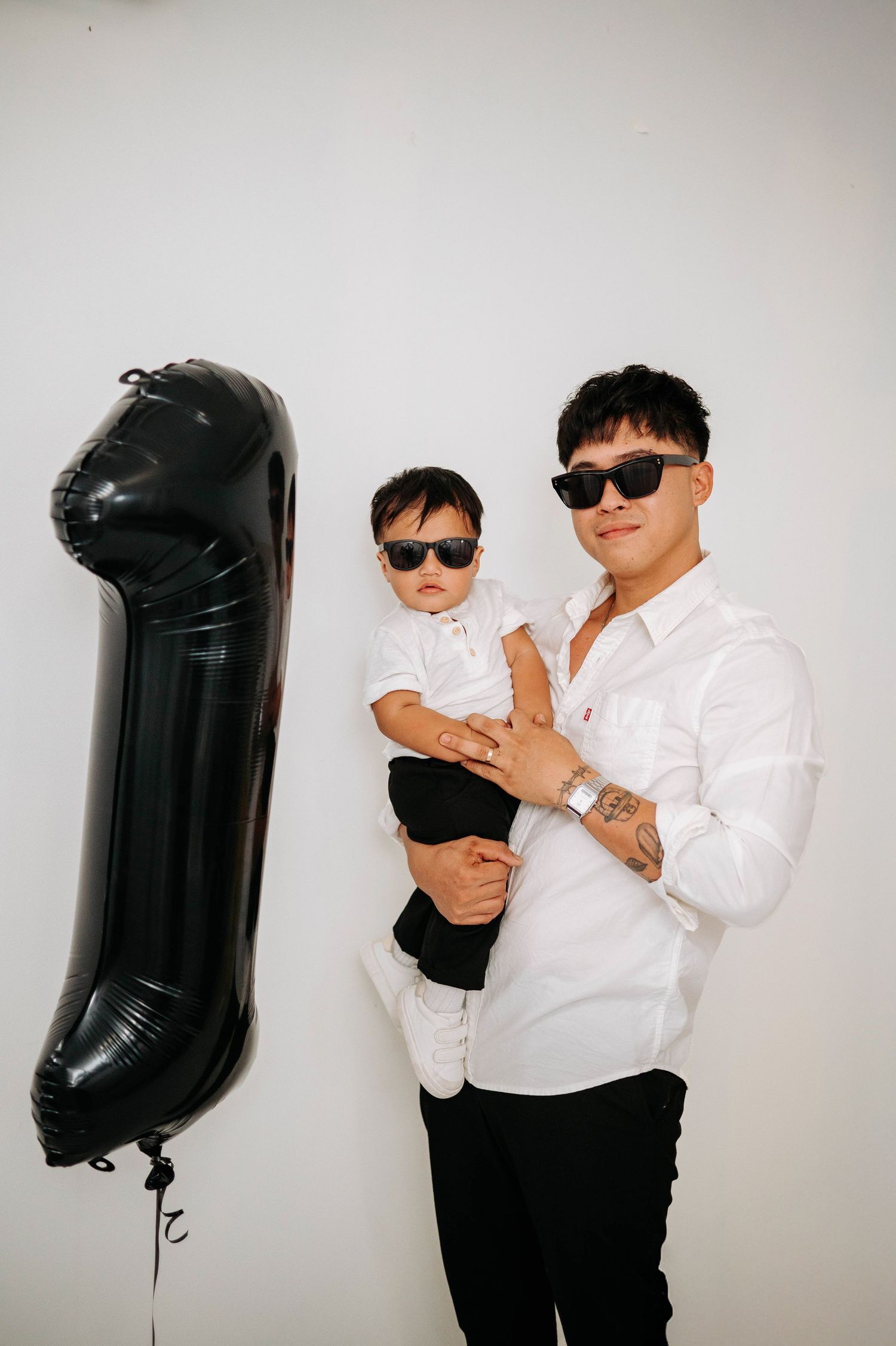 Contemporary monochromatic photo with two figures in matching white shirts and sunglasses posing next to a black balloon.