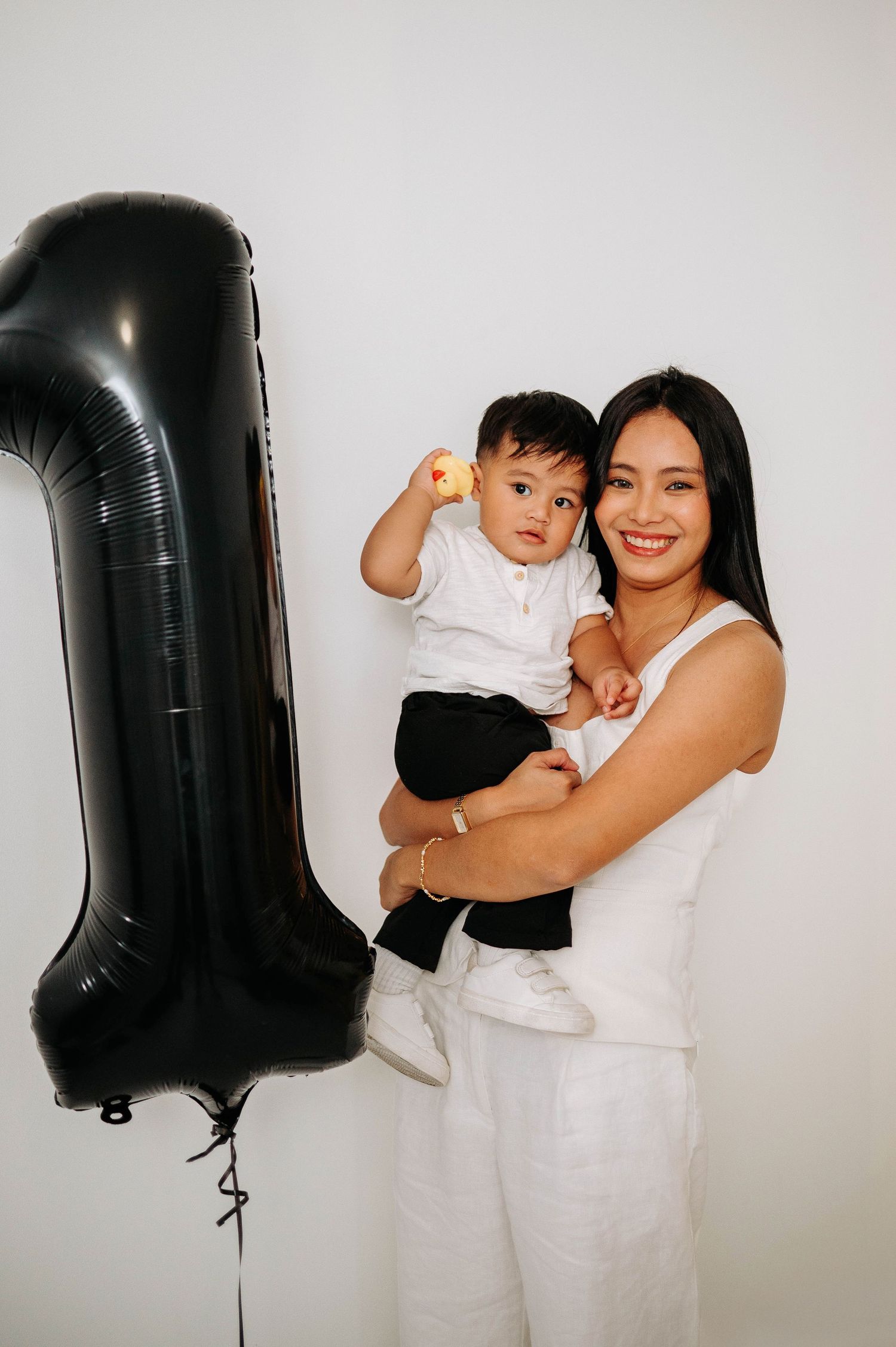 Woman in white holding toddler next to large black number one balloon against white background.